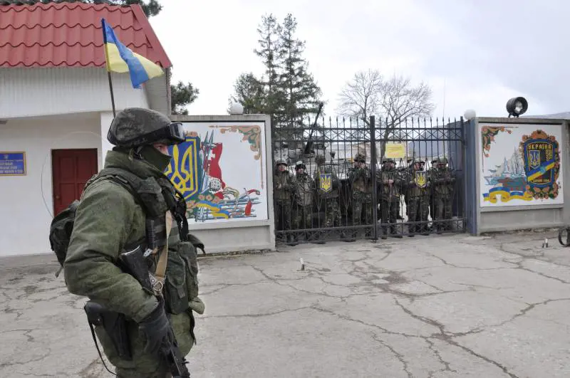 In primo piano soldato russo che circonda la base e soldati ucriaini dietro che presidiano il cancello d'ingresso